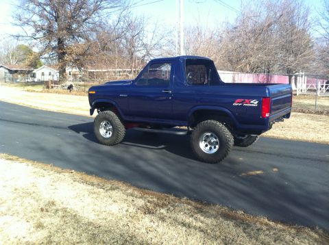 1982 Ford Bronco - A lifted blue Ford Bronco with off-road tires drives down a road in a rural setting.