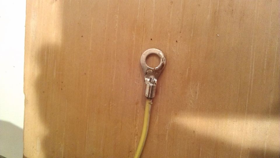 Electrical ring terminal crimped onto a yellow wire, against a wood-grain background