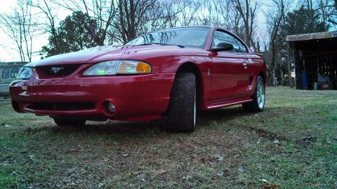 1997 Ford Mustang - A red 1997 Ford Mustang coupe is parked on a grassy area with trees in the background.