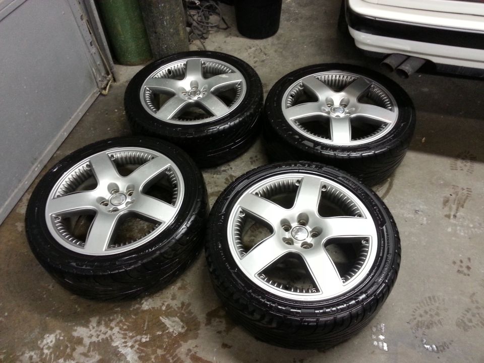 Four silver alloy wheels with black tires stacked in a garage. A white car's rear bumper and exhaust pipes are visible to the right