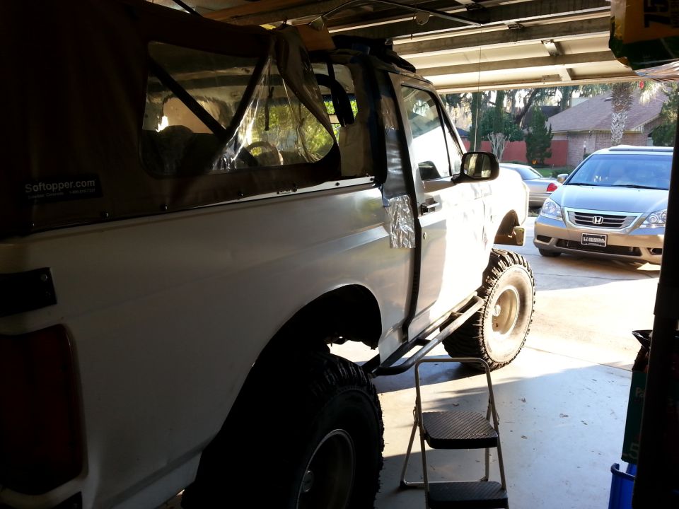 Side view of a white Ford Bronco with a brown soft top and large tires, parked in a garage. Another car is visible in the background