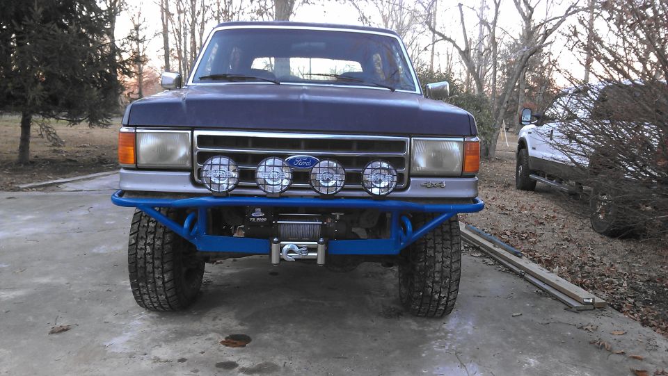 Front view of a dark blue Ford truck with a blue custom bumper, winch, and four spotlights. Another white truck is parked nearby