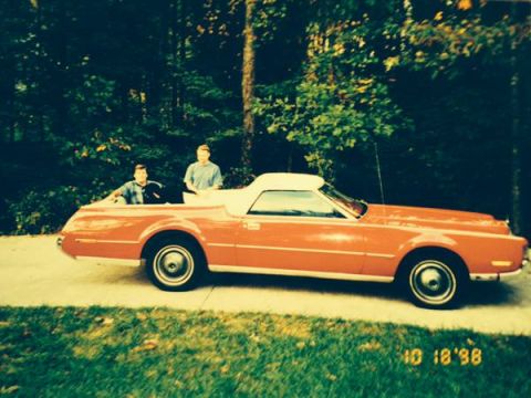 1973 Ford Ranchero - A side view of an orange convertible coupe with two occupants, set against a backdrop of trees.