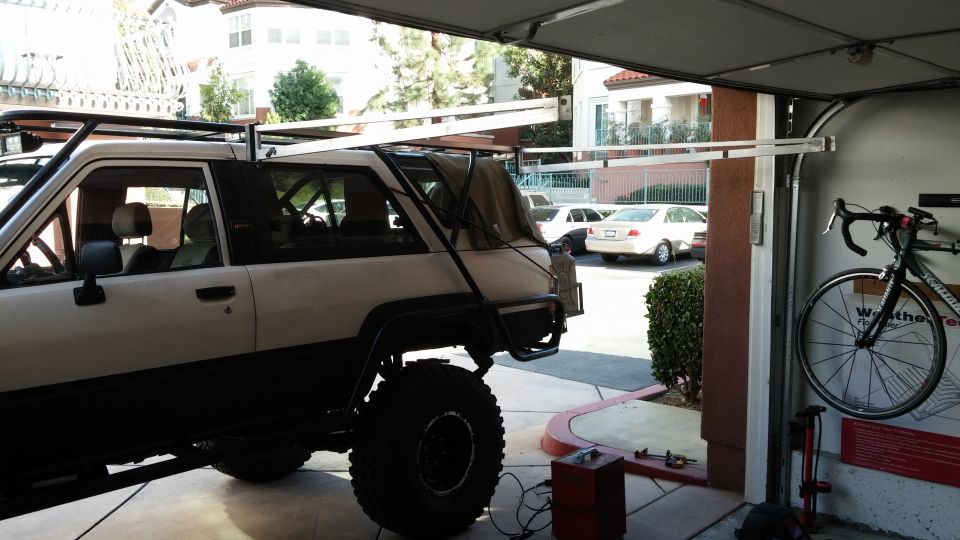 White SUV with large tires and roof rack parked in a garage, other cars visible outside