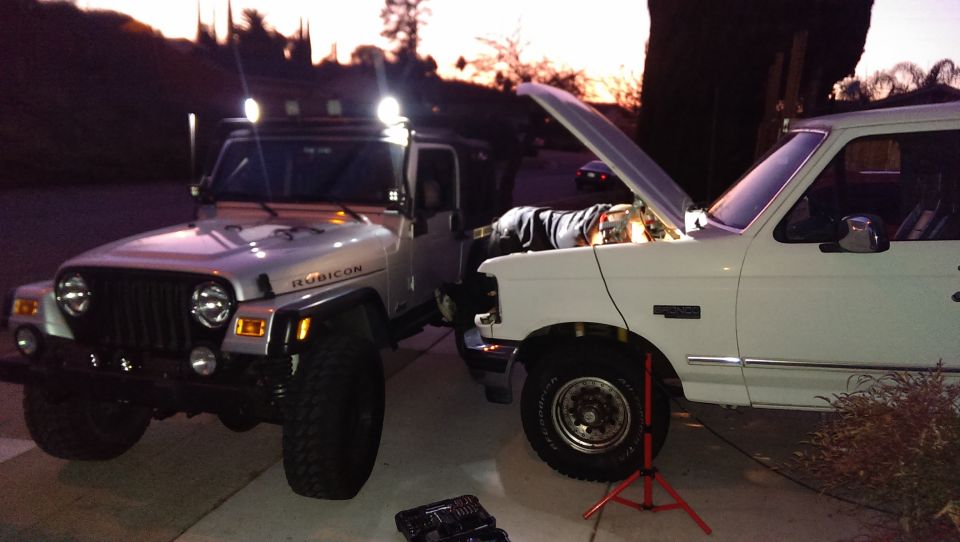 Jeep Wrangler and white Ford truck with hood open, parked outside at dusk