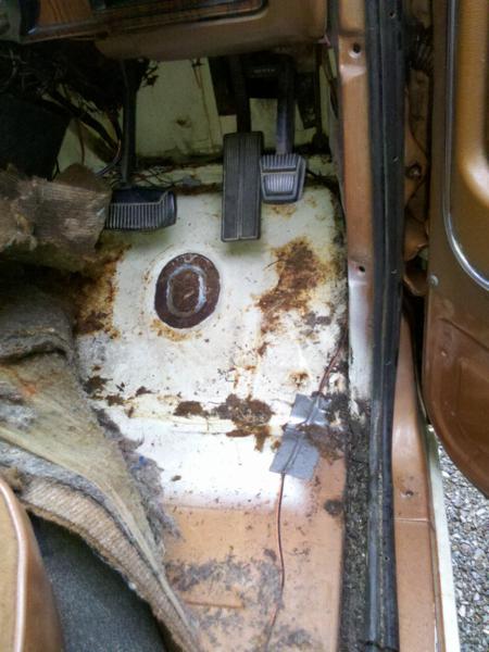 1982 Ford Bronco - The interior floor of a vehicle, pedals, significant rust, and worn carpeting.