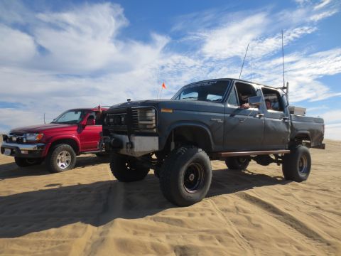 1989 Ford Bronco - Two off-road vehicles are parked on a sandy terrain with a bright orange flag visible in the distance.