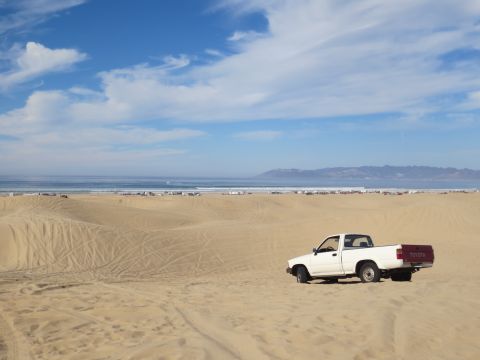 1989 Ford Bronco - A white Toyota pickup truck with a red tailgate sits on sand dunes with the ocean and distant mountains in the background.