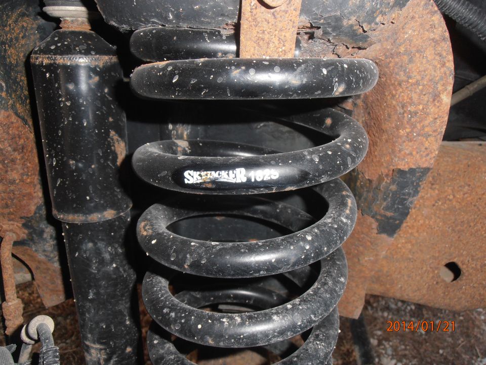 Close-up of a black coil spring with white lettering "SKYJACKER 1025" and a black shock absorber, showing rust on surrounding metal parts