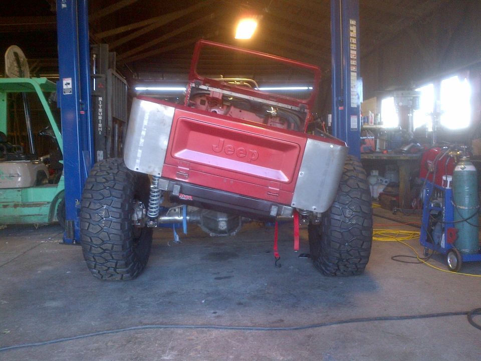 Red Jeep CJ with custom rear fenders and large tires, lifted on a car hoist in a garage