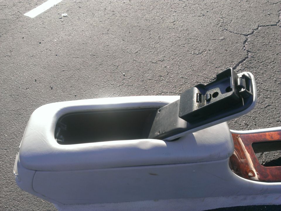 Gray leather center console with lid open, removed from car, sitting on asphalt with woodgrain trim piece nearby