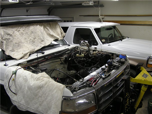 White Ford truck with hood removed and fenders covered, engine bay exposed in a garage