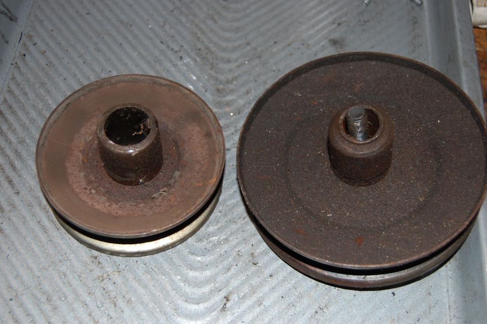 Two rusty metal pulleys of different sizes on a gray surface. The pulley on the left is smaller and has a white band around its outer edge. The pulley on the right is larger and entirely dark brown/black