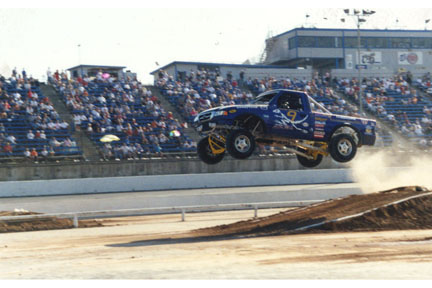 1993 Ford Taurus - A blue monster truck soars through the air during a jump, with spectators filling the grandstands behind it.