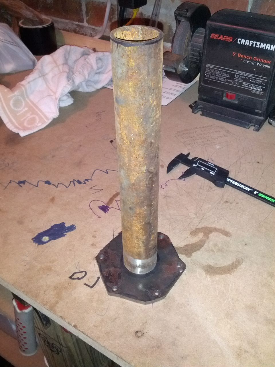 Rusted metal pipe mounted on a hexagonal metal plate, with a digital caliper and a bench grinder in the background