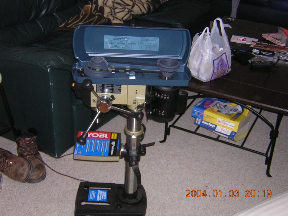 Drill press with a Ryobi box on it. Boots and a coffee table with boxes are nearby