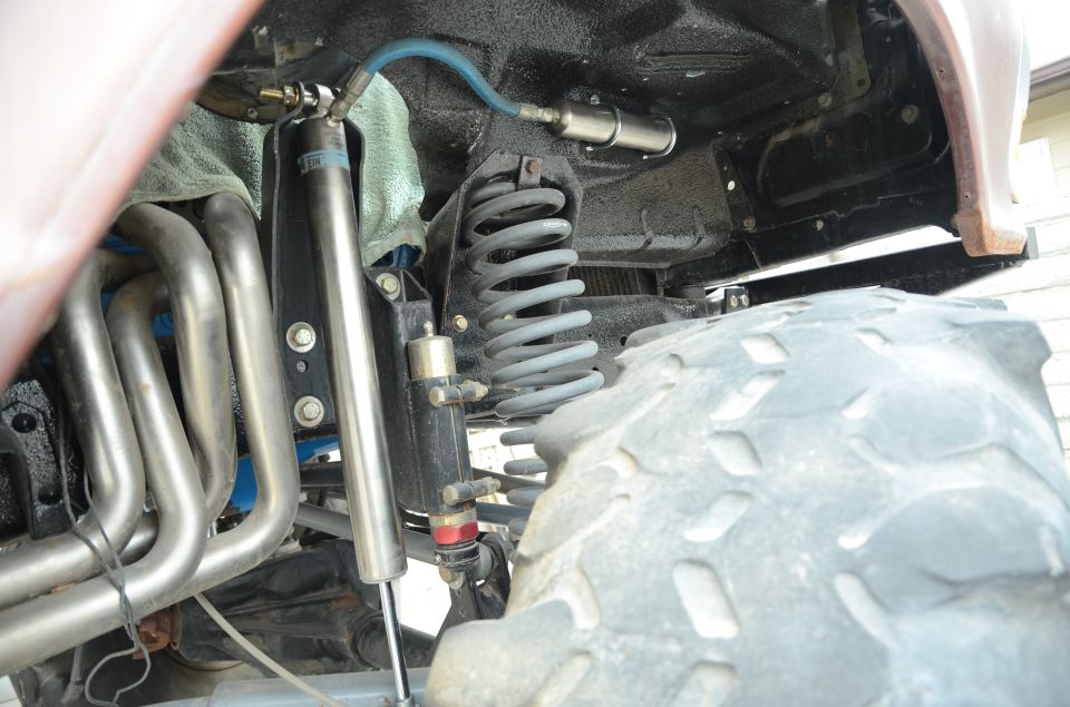 Close-up of off-road vehicle's front suspension with large coil spring, shock absorber, and exhaust headers. Large tire tread visible
