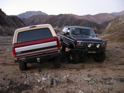 1985 Ford Bronco - Two modified Ford Broncos are parked on a dirt trail with mountains in the background.