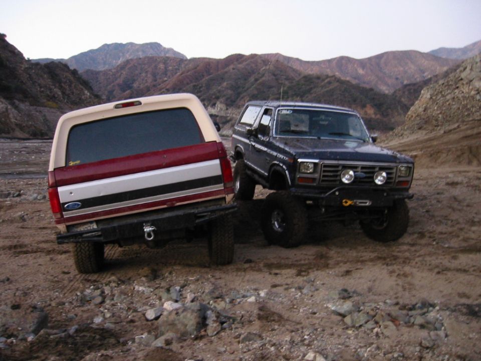 Rear of two-tone Bronco next to dark blue lifted Bronco in desert with mountains