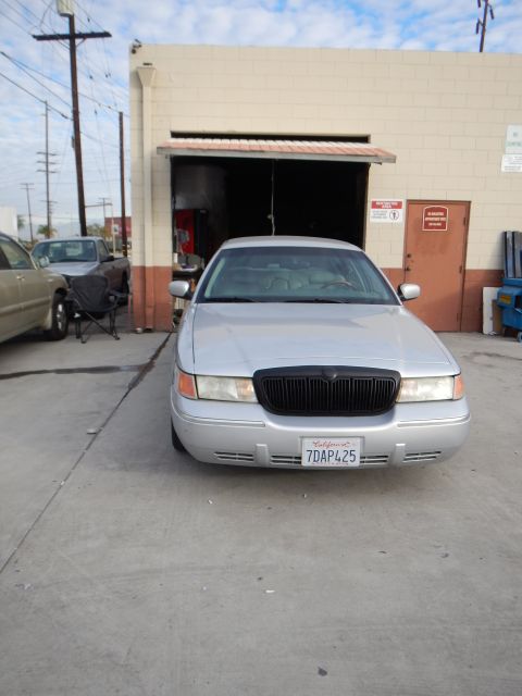 2003 Ford Crown Victoria - silver four-door sedan with California plate 7DAP425 parked head-on outside an industrial auto shop bay.