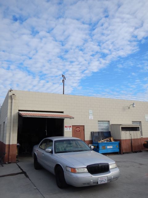 2003 Ford Crown Victoria - silver four-door sedan parked beside an open bay of a beige industrial auto shop under a bright streaky-cloud sky.