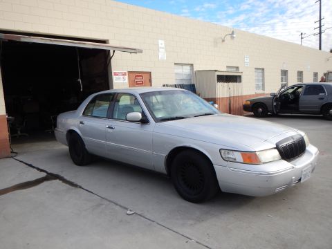 2003 Ford Crown Victoria - silver four-door sedan fitted with black steel wheels sits at an angle outside an industrial auto shop with open bays.