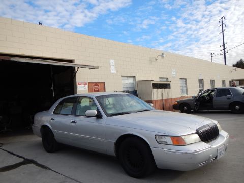 2003 Ford Crown Victoria - silver four-door sedan with black wheels parked along a long beige industrial building with open shop bays.