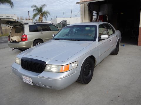 2003 Ford Crown Victoria - silver Mercury four-door sedan with blacked-out grille and wheels, plate 7DAP425, parked outside a shop with a gold SUV behind.