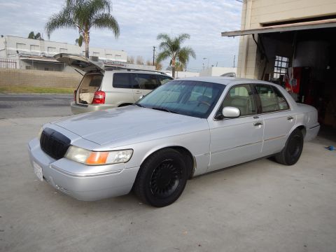 2003 Ford Crown Victoria - silver Mercury four-door sedan with blacked-out grille and black wheels parked outside an auto shop, gold SUV and palm trees behind.