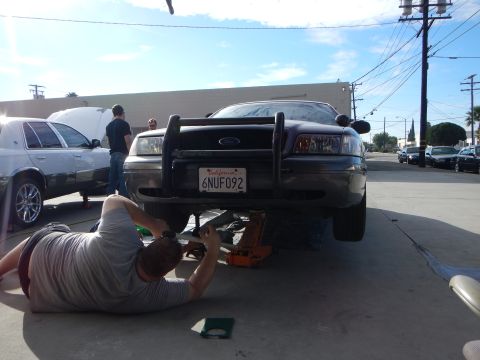 2003 Ford Crown Victoria - dark Ford sedan fitted with a tubular brush guard, plate 6NUF092, sits on a floor jack while a person lies beneath it working.