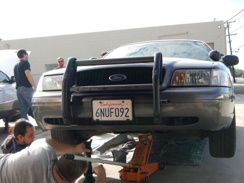 2003 Ford Crown Victoria - Close front view of a dark Ford sedan with a black tubular push bar and California plate 6NUF092, raised on a floor jack.