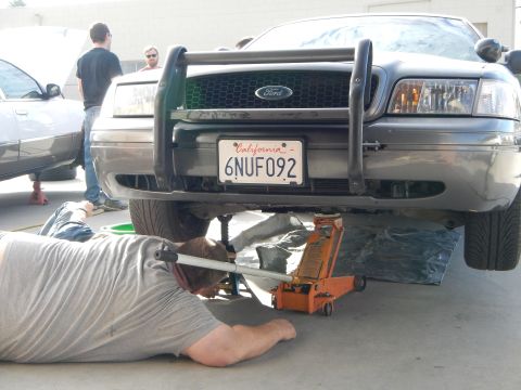 2003 Ford Crown Victoria - person lies on the ground working under the front of a dark Ford sedan with push bar, plate 6NUF092, raised on an orange floor jack.