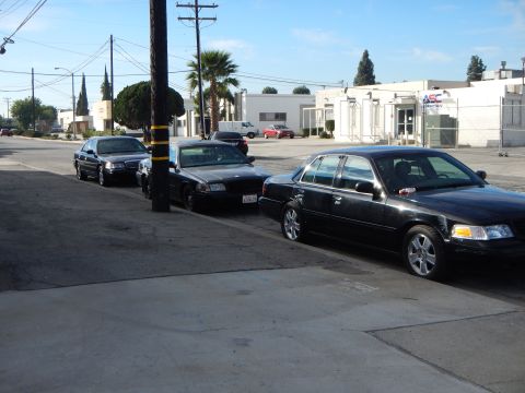 2003 Ford Crown Victoria - Several dark-colored four-door sedans parked nose-to-tail along a curb on an industrial street under a partly cloudy sky.
