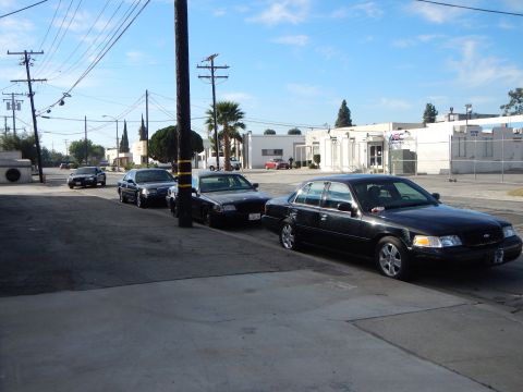 2003 Ford Crown Victoria - line of black four-door sedans parked along the curb of a sunny industrial street, palm trees and warehouses behind.
