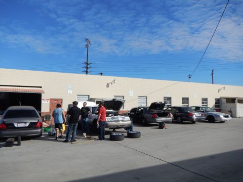 2003 Ford Crown Victoria - Several sedans with hoods and trunks open are serviced by people in an open lot outside a long beige industrial building.
