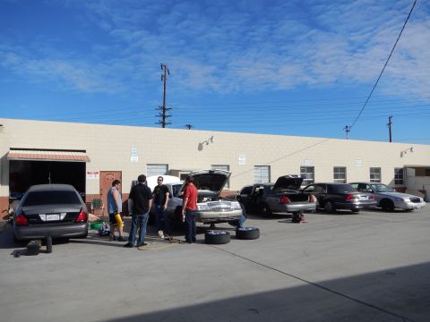 2003 Ford Crown Victoria - group of people work on a row of sedans with raised hoods and loose tires in the lot of a beige industrial auto shop.