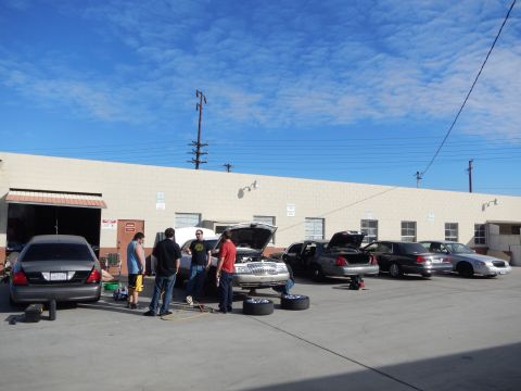 2003 Ford Crown Victoria - People gather around a row of sedans with open hoods and tires on the ground in the lot of a beige industrial shop under blue sky.
