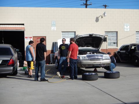 2003 Ford Crown Victoria - Several people stand and crouch around a silver sedan with its hood up and two tires on the ground outside an auto shop.