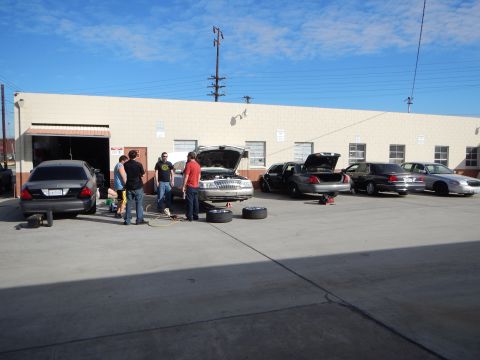 2003 Ford Crown Victoria - group of people work on a row of parked sedans with hoods and trunks open and loose tires in a beige industrial shop lot.