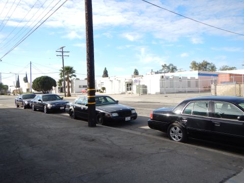 2003 Ford Crown Victoria - Black four-door sedans parked nose-to-tail along a curb on a sunny industrial street with warehouses across the road.