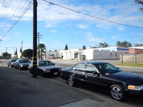 2003 Ford Crown Victoria - line of black four-door sedans parked along the curb of an industrial street under a bright blue sky with scattered clouds.