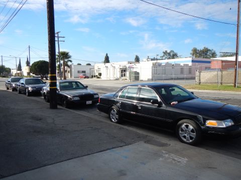 2003 Ford Crown Victoria - Several black four-door sedans parked nose-to-tail along a curb on a sunny industrial street lined with warehouses and palm trees.