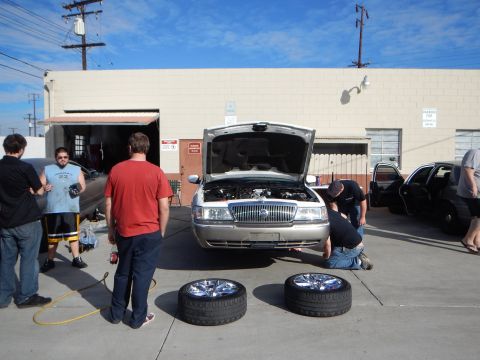 2003 Ford Crown Victoria - group of people stand and crouch around a silver Mercury sedan with its hood open while two chrome-wheel tires rest on the pavement.