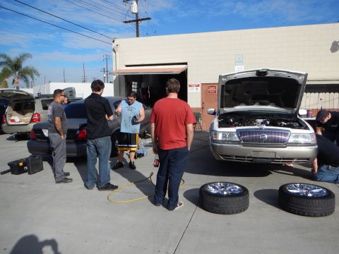 2003 Ford Crown Victoria - Several men gather around a silver sedan with its hood raised in a sunny shop lot, with two alloy wheels and tires lying on the pavement.