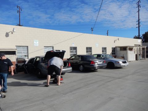 2003 Ford Crown Victoria - Three full-size sedans line up beside a tan industrial building while a man leans into the open trunk of the nearest car.