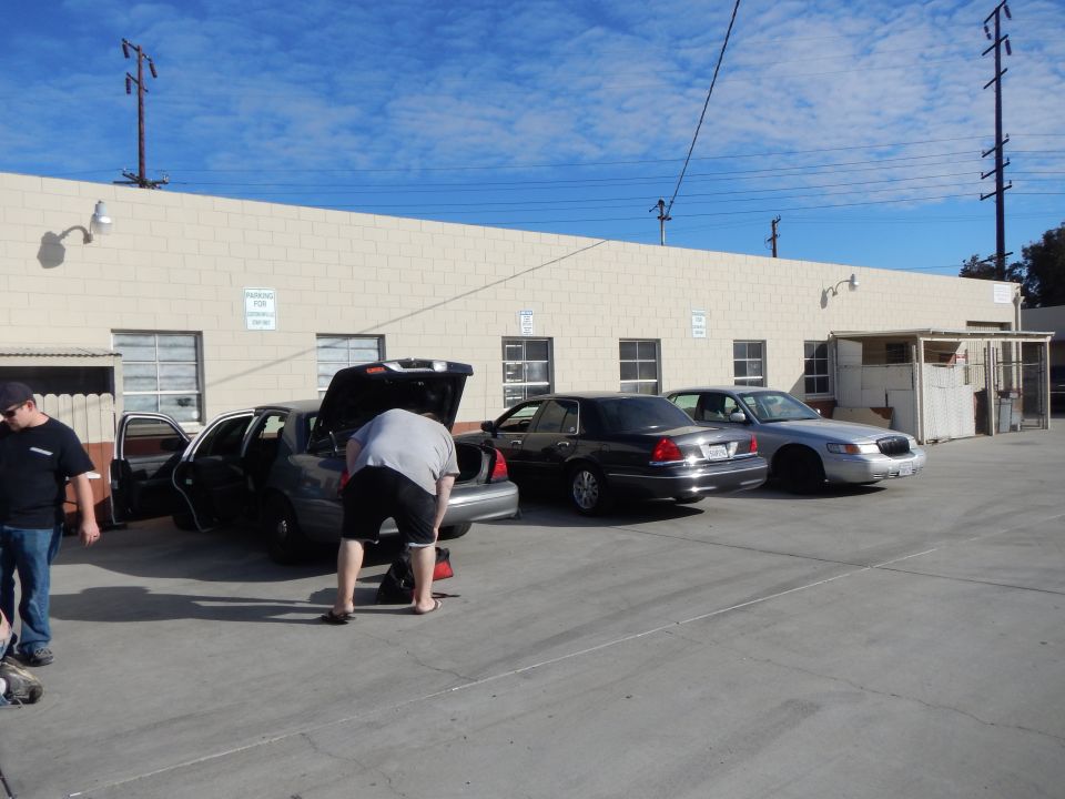 Three full-size sedans parked along a tan industrial building, a man leaning into an open trunk