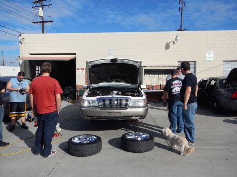 2003 Ford Crown Victoria - silver sedan sits with its hood open in a shop lot as men stand around it, two chrome wheels on the pavement and a small dog nearby.
