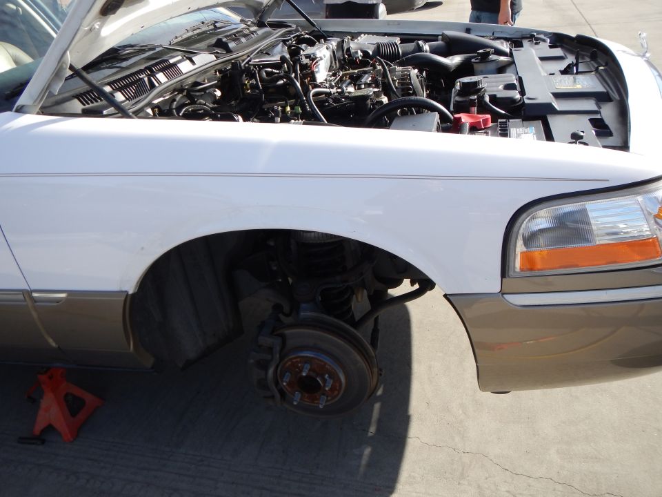 Close-up of a white and tan car's front corner with fender off, engine and brake hub exposed