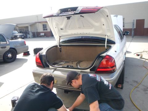 2003 Ford Crown Victoria - Two men crouch at the rear bumper of a silver sedan with its trunk open, working on components inside a sunny shop lot.