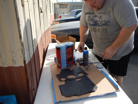 2003 Ford Crown Victoria - man in a Vegas t-shirt works on a black metal panel taped to cardboard on a folding table, with spray cans beside him.